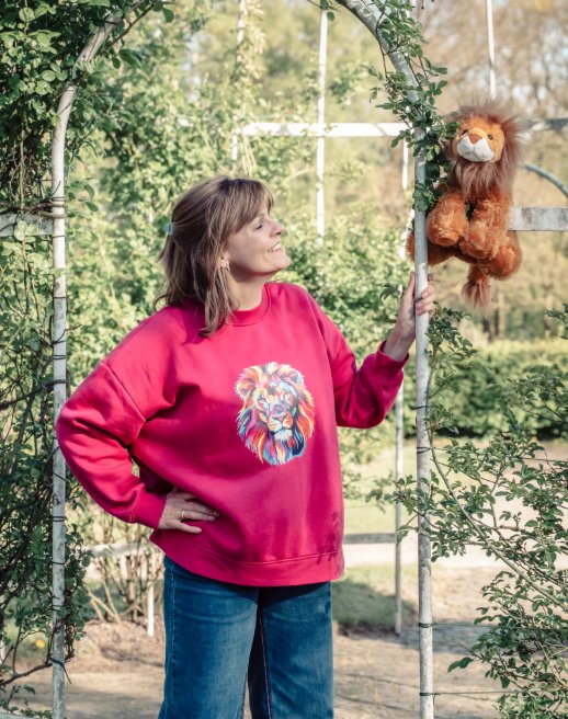 Nathalie Barth in einem pinkfarbenen Pullover mit Löwenmotiv in einem Rosengarten, neben ihr ein Stoff-Löwe am Rosenbogen